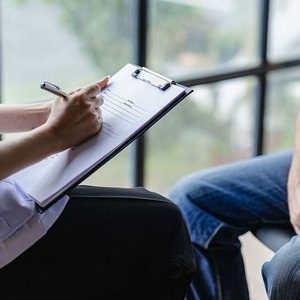 Mensch mit Klemmbrett und Stift sitzt einer anderen Person gegenüber